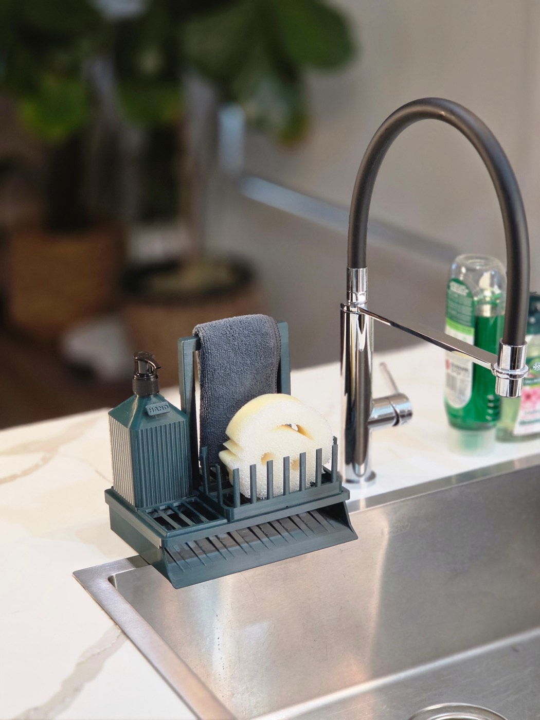 The Essentials Station on a kitchen sink, holding a soap pump, a cloth and a sponge, draining into the bowl.